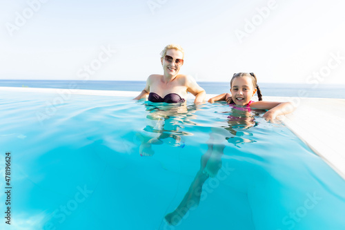 Young pretty woman and cute little girl in summer bikini and sunglasses posing together sitting near the pool in hotel. Copy space.