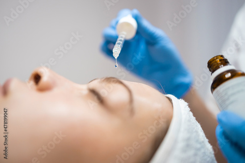 Close-up of a beautician's hand with a pipette dripping nourishing oil serum to the skin of a young woman's face