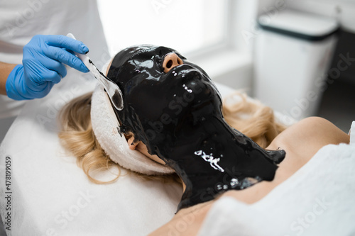 Beautician apply a black alginate mask with a spatula to the face of a young woman. Cosmetic jelly mask. Facial skin care photo