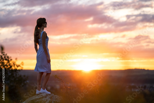 A young woman in summer dress standing outdoors enjoying view of bright yellow sunset © bilanol