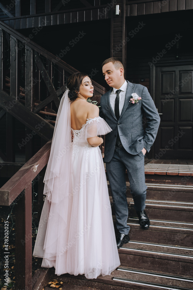 Stylish groom and a beautiful brunette bride in a white dress with a bouquet in their hands are standing and hugging on the background of a wooden house with a ladder. Wedding portrait. Stylish groom and a beautiful brunette bride in a white dress with a bouquet in their hands are standing and hugging on the background of a wooden house with a ladder. Wedding portrait.