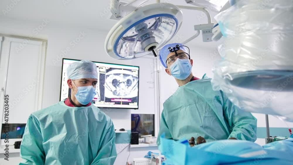 Two neurosurgeons stand over the patient on a table. Doctors apply instruments to perform operation. Professionals look at the screen. Two neurosurgeons stand over the patient on a table. Doctors apply instruments to perform operation. Professionals look at the screen.