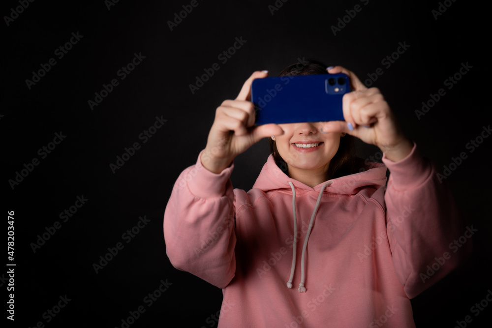 Cute young brunette in a pink hoodie uses a smartphone and laughs, communication without borders, concept portrait on a black background Cute young brunette in a pink hoodie uses a smartphone and laughs, communication without borders, concept portrait on a black background