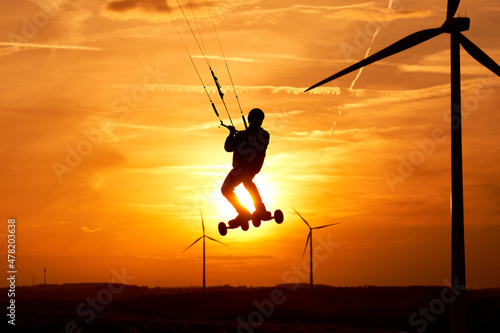 Kite land boarder jumps in front of setting sun. 1 man at an extreme sport art in action with a kiteboard. Landscape with wind turbines as silhouette. Front view.