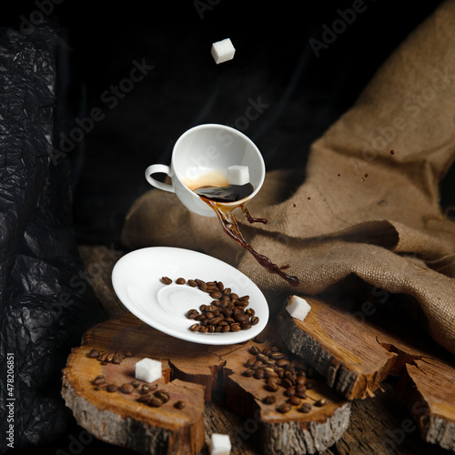 refined sugar cubes fall into a white cup of coffee, coffee is spilled on a saucer where roasted coffee beans lie