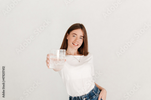 Selective focus portrait of lively brunette woman dressed in white t-shirt and jeans, offering a glass of pure water to the camera over white background