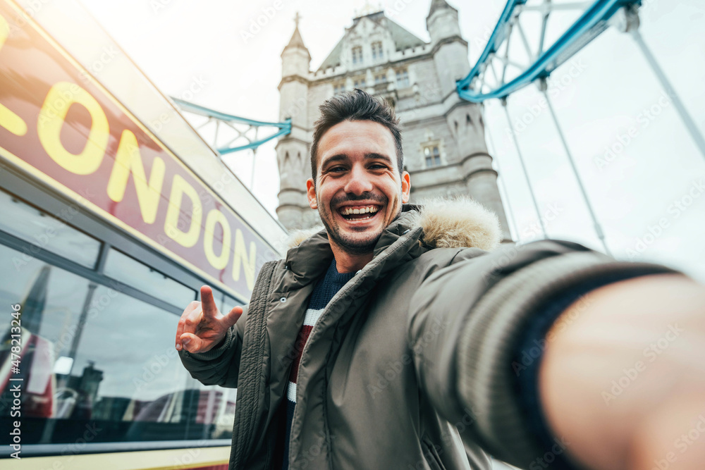 Smiling man taking selfie portrait during travel in London, England - Young tourist male taking memory pic with iconic england landmark - Happy people wandering around Europe concept - obrazy, fototapety, plakaty 