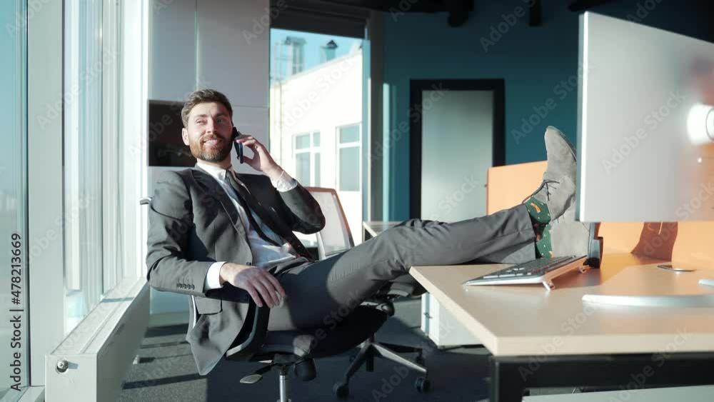 Handsome Business man boss in suit formalwear talking on mobile phone telling about his achievement, sitting at the modern office with feet on the desk top. Happy cheerful male employee entrepreneur Handsome Business man boss in suit formalwear talking on mobile phone telling about his achievement, sitting at the modern office with feet on the desk top. Happy cheerful male employee entrepreneur