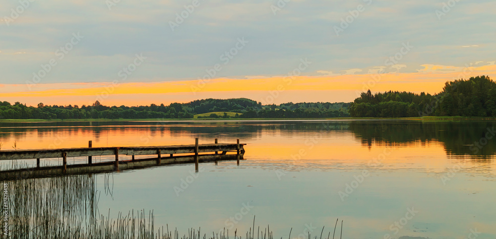 A lake with the reflection of clouds and trees at sunset. A long wooden footbridge leads into the water. A drawn illustration of a lake