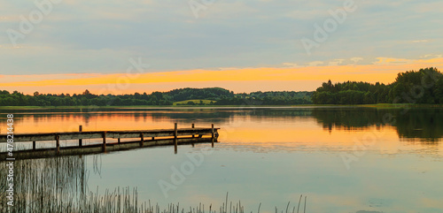 Fototapeta Naklejka Na Ścianę i Meble -  A lake with the reflection of clouds and trees at sunset. A long wooden footbridge leads into the water. A drawn illustration of a lake
