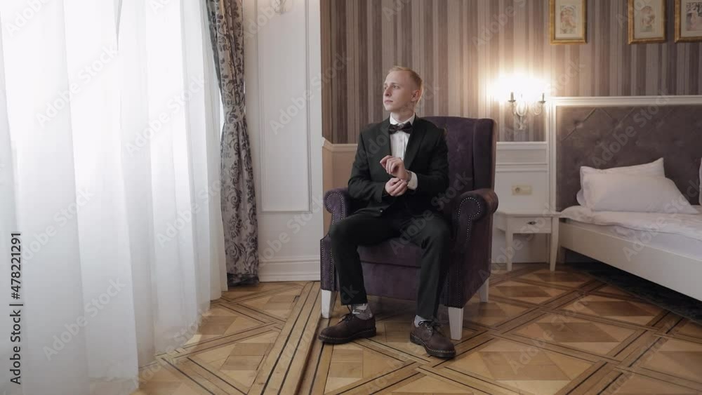 Man in suit put on old-fashioned wrist watch, sitting on chair. Groom dresses. Handsome male businessman waiting for meeting. Hand with clock close-up. Waits. Looks at clock, time. Wristwatch on arm Man in suit put on old-fashioned wrist watch, sitting on chair. Groom dresses. Handsome male businessman waiting for meeting. Hand with clock close-up. Waits. Looks at clock, time. Wristwatch on arm
