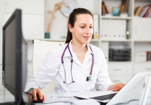 Confident young female doctor prints an outpatient patient card on a computer in the office, sitting at her workplace