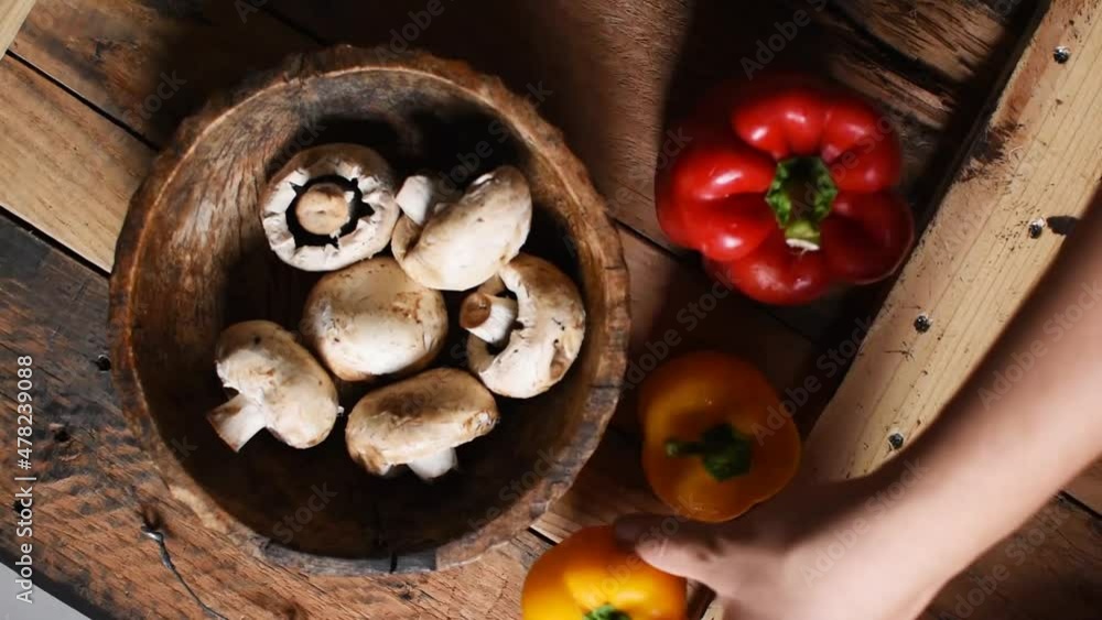 hand putting two yellow pepper next to a red pepper and wooden bowl with white mushrooms inside on rustic wooden texture as background-flat lay, steady cam hand putting two yellow pepper next to a red pepper and wooden bowl with white mushrooms inside on rustic wooden texture as background-flat lay, steady cam