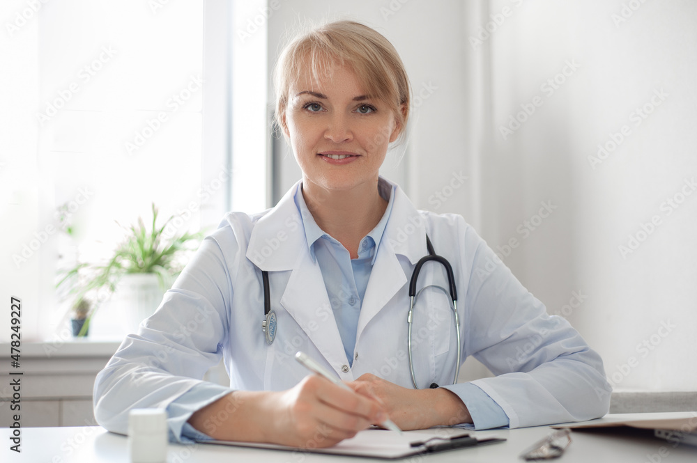 Portrait of a beautiful middle aged female doctor wears white coat stethoscope on shoulders looking at camera. Online counseling and therapy concept Horizontal photo. E-health. Woman smiling. Portrait of a beautiful middle aged female doctor wears white coat stethoscope on shoulders looking at camera. Online counseling and therapy concept Horizontal photo. E-health. Woman smiling.