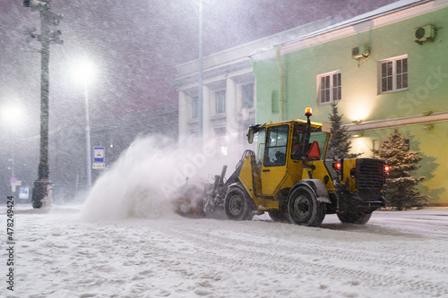 Tractor with rotary sweeper clearing city street at night after heavy snowfall. Clearing city by municipalities during winter season. Professional equipment for snow removal. Winter road maintenance