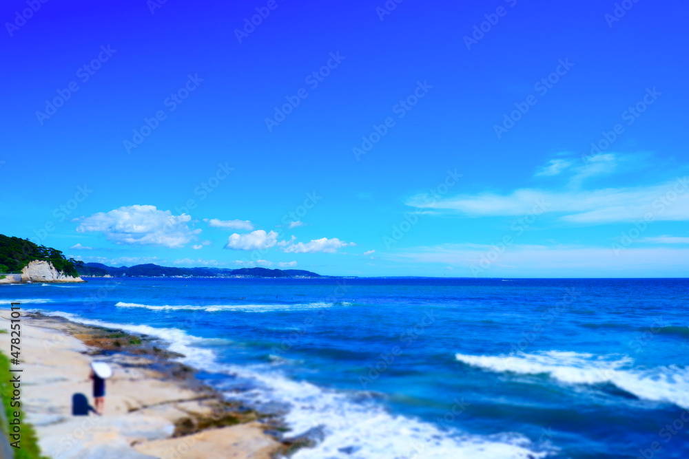 よく晴れた湘南の海と、サーフボードを持って波打ち際を歩くサーファー