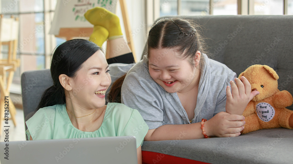 Fototapeta premium Portrait shot of Asian lovely mother sitting on floor and young chubby down syndrome autistic autism little daughter lay down on cozy sofa smiling look at camera watching cartoon from laptop computer
