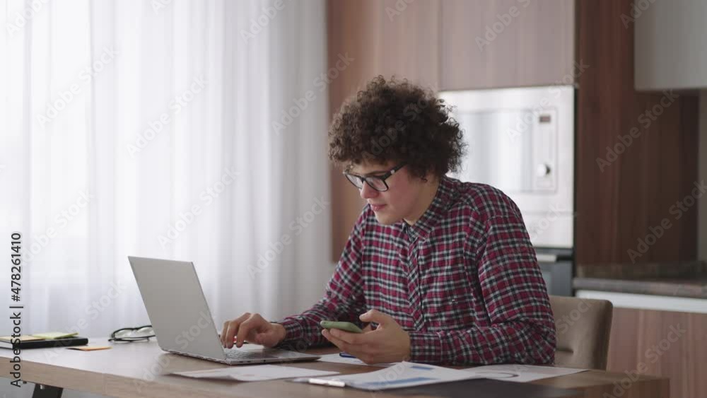 Young man with smartphone in his hands. Modern businessman or student at home office. Freelancer at work. Young student man study at home with laptop and uses a smartphone Young man with smartphone in his hands. Modern businessman or student at home office. Freelancer at work. Young student man study at home with laptop and uses a smartphone