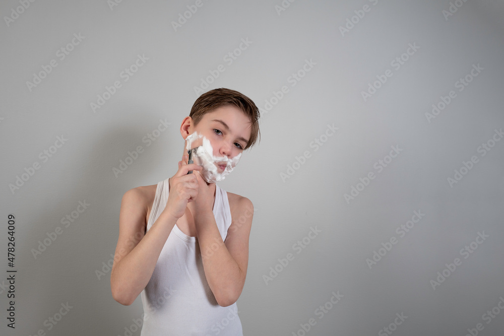 Cute boy with shaving foam and razor playfully posing against the background of a gray wall.