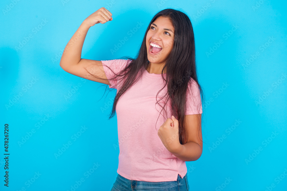 Attractive Young hispanic girl wearing pink T-shirt over blue background celebrating a victory punching the air with his fists and a beaming toothy smile Attractive Young hispanic girl wearing pink T-shirt over blue background celebrating a victory punching the air with his fists and a beaming toothy smile
