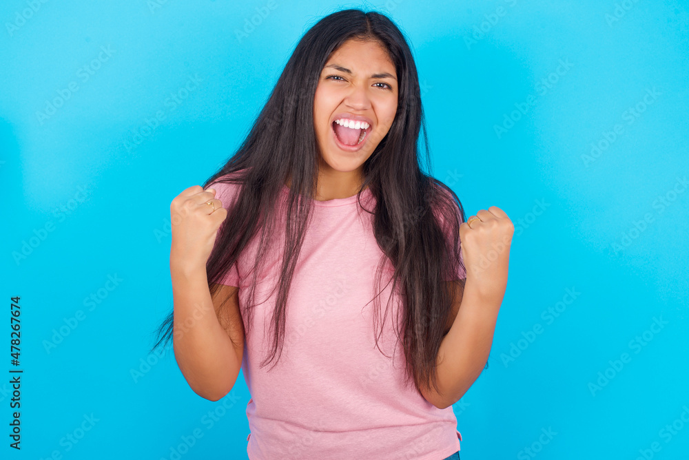 Portrait of Young hispanic girl wearing pink T-shirt over blue background looks with excitement at camera, keeps hands raised over head, notices something unexpected reacts on sudden news. Portrait of Young hispanic girl wearing pink T-shirt over blue background looks with excitement at camera, keeps hands raised over head, notices something unexpected reacts on sudden news.