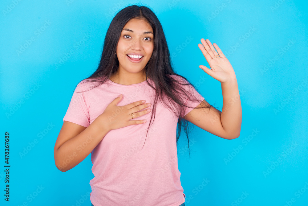 I swear, promise you not regret. Portrait of sincere Young hispanic girl wearing pink T-shirt over blue wall raising one arm and hold hand on heart as give oath, telling truth, want you to believe.