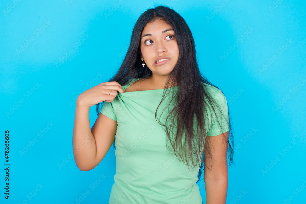 Young hispanic girl wearing green T-shirt over blue background stressed, anxious, tired and frustrated, pulling shirt neck, looking frustrated with problem Young hispanic girl wearing green T-shirt over blue background stressed, anxious, tired and frustrated, pulling shirt neck, looking frustrated with problem