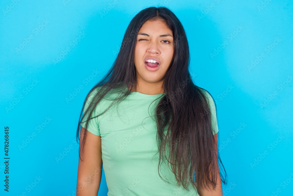 Young hispanic girl wearing green T-shirt over blue background yawns with opened mouth stands. Daily morning routine Young hispanic girl wearing green T-shirt over blue background yawns with opened mouth stands. Daily morning routine