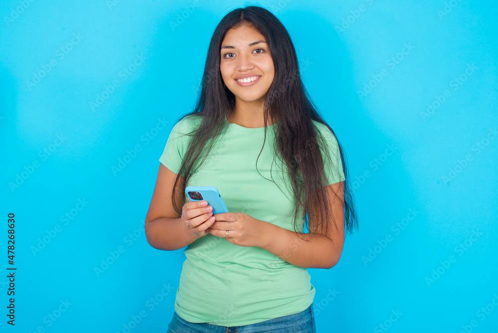 Young hispanic girl wearing green T-shirt over blue background Mock up copy space. Using mobile phone, typing sms message Young hispanic girl wearing green T-shirt over blue background Mock up copy space. Using mobile phone, typing sms message