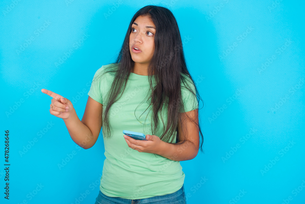Stunned Young hispanic girl wearing green T-shirt over blue background points sideways right copy space, recommends product, sees astonishing thing