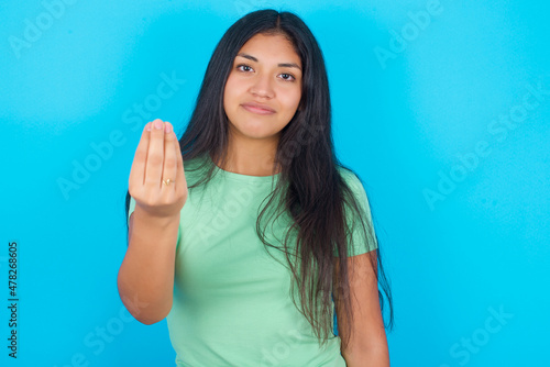 Young hispanic girl wearing green T-shirt over blue background Doing Italian gesture with hand and fingers confident expression