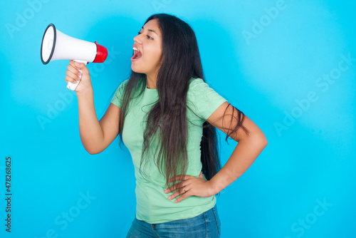 Funny Young hispanic girl wearing green T-shirt over blue background People sincere emotions lifestyle concept. Mock up copy space. Screaming in megaphone.
