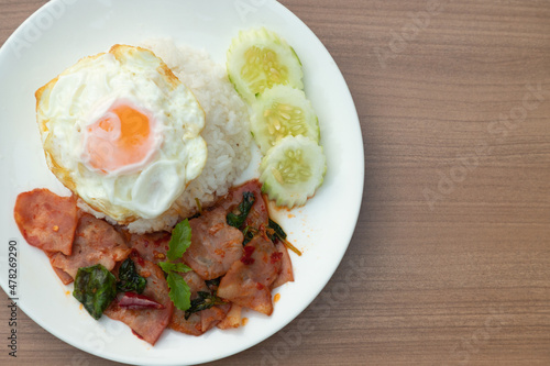 Closeup of Thai traditional fusion food, Flavorful appetizing food as Spicy bacon with basil leaves, fried egg in white modern plate on clean minimal wooden table background and texture, copy space.
