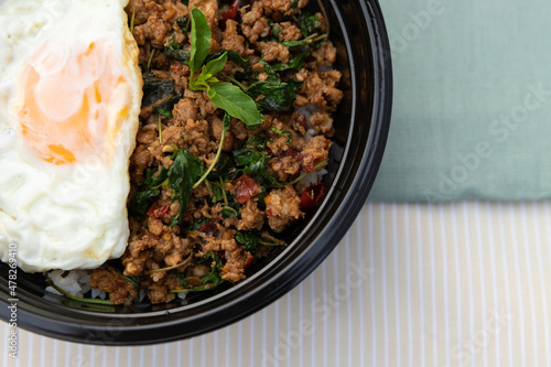 Closeup of Thai traditional food, Flavorful appetizing food as Spicy fried pork with basil leaves, fried egg and cucumber in black modern bowl on clean minimal cream and green table cloth background