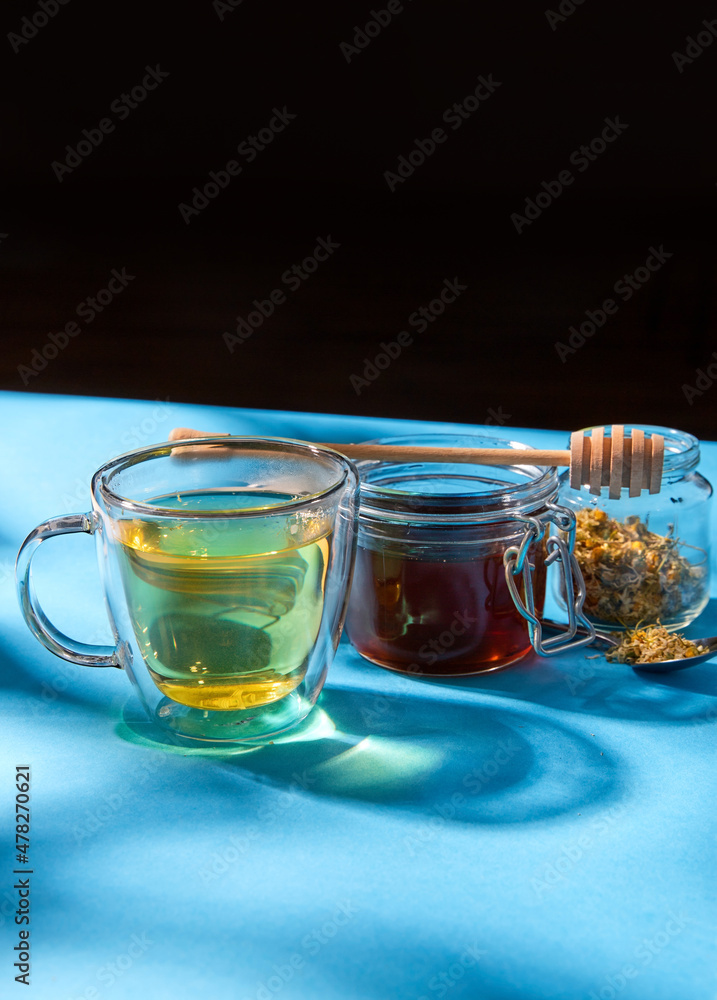 folk medicine and health concept - close up of cup of herbal tea, honey with dipper and dry chamomile flowers in glass jar on blue background folk medicine and health concept - close up of cup of herbal tea, honey with dipper and dry chamomile flowers in glass jar on blue background