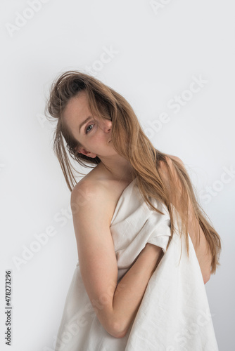 Young nude woman with long hair is covered with white sheet. Portrait of beautiful caucasian girl in the morning. White background