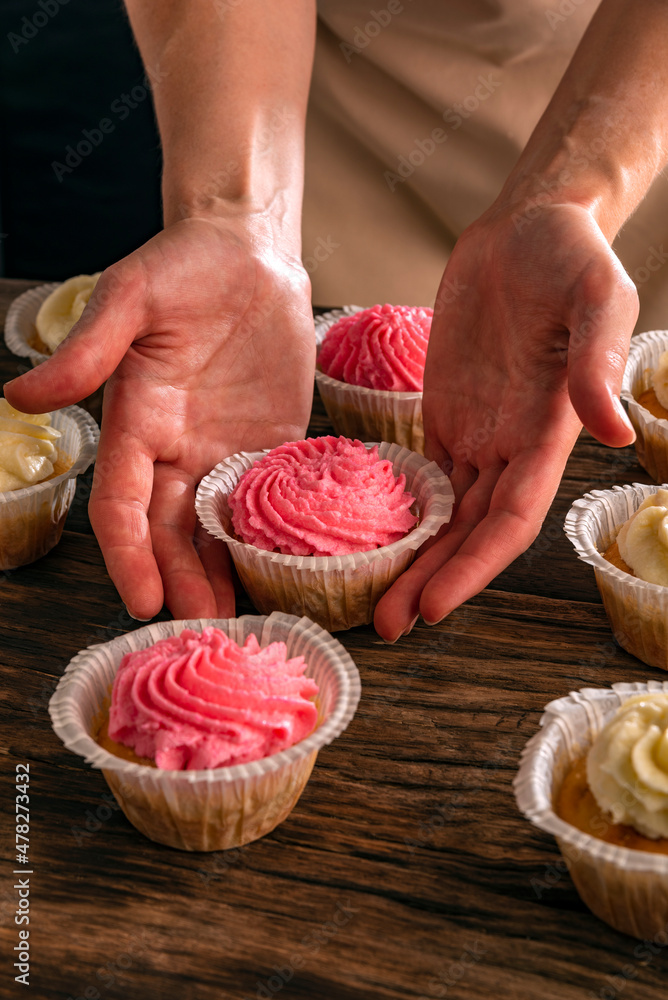 Female hands keeps vanilla cupcake with pink cream. Just cooked cakes on wooden table. Vertical frame.