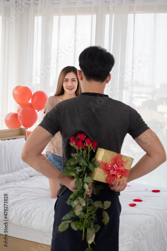 The girlfriend is waiting for something hidden behind her boyfriend on the bed in the bedroom decorated with red heart balloons on Valentine's Day.