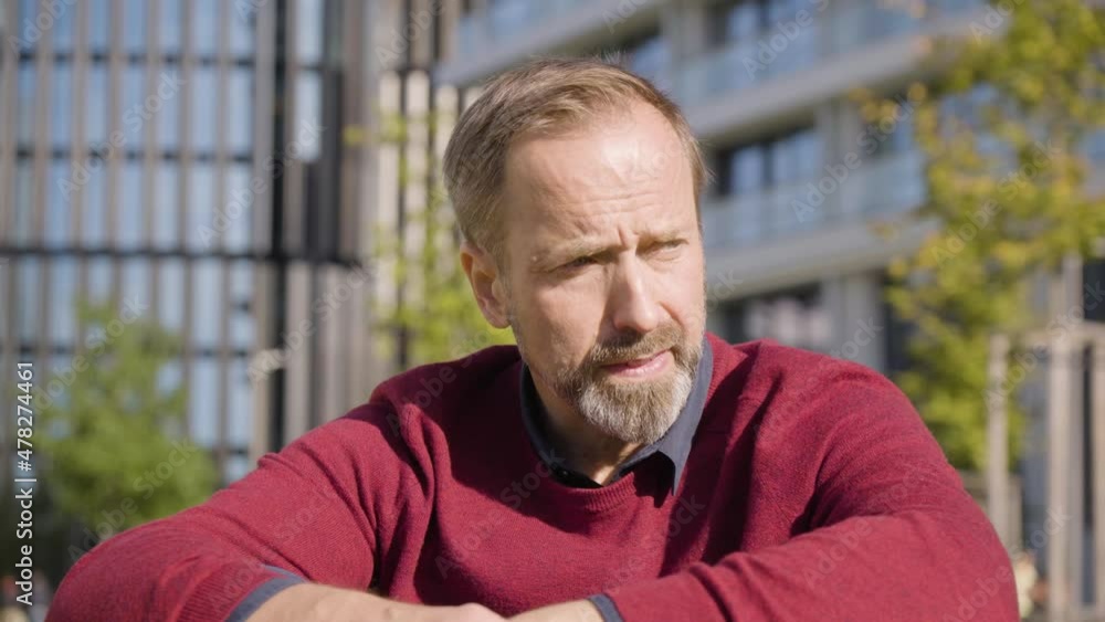 A middle-aged handsome Caucasian man thinks about something as he sits in a street in an urban area - office buildings in the blurry background - closeup A middle-aged handsome Caucasian man thinks about something as he sits in a street in an urban area - office buildings in the blurry background - closeup
