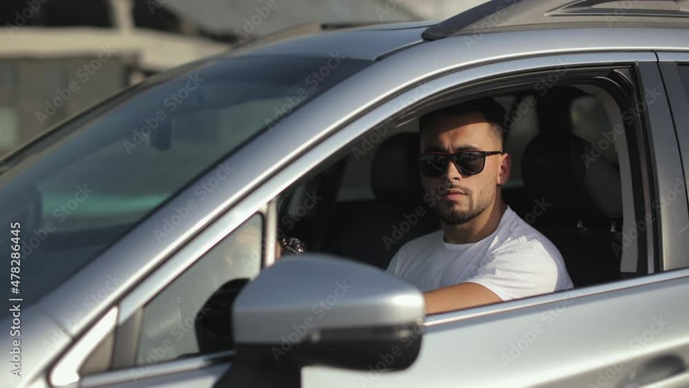 The man is sitting in the car in the driver's seat. He is turning his head and looking at the camera. He's wearing sunglasses. 4K The man is sitting in the car in the driver's seat. He is turning his head and looking at the camera. He's wearing sunglasses. 4K