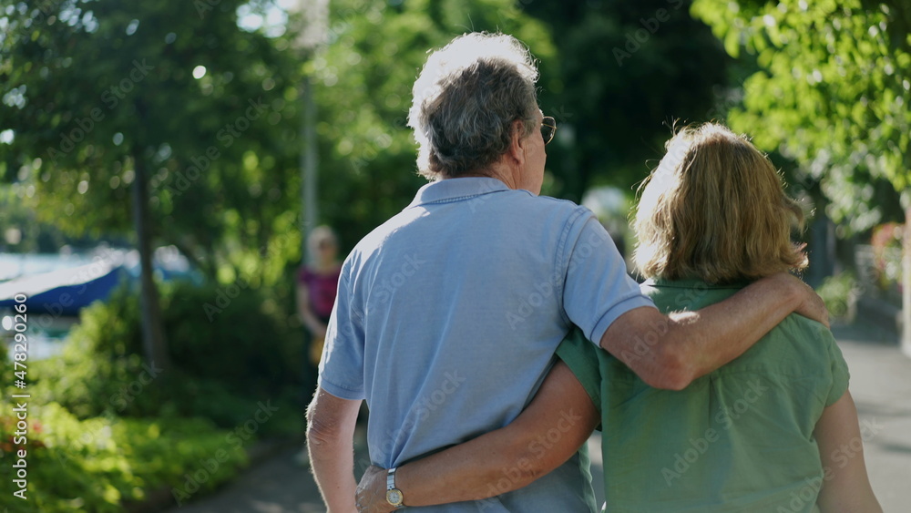 Romantic senior husband putting arm around wife, back view of older couple walking together in green path Romantic senior husband putting arm around wife, back view of older couple walking together in green path