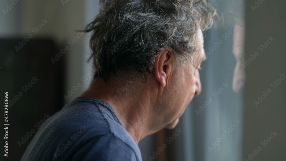Fototapeta premium Pensive senior man standing by window looking outside. Thoughtful old person