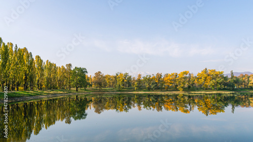 Autumn landscape with a view of the lake, trees and mountains. Reflection of yellowed foliage of trees in the water. Blue sky and mountains in the distance.