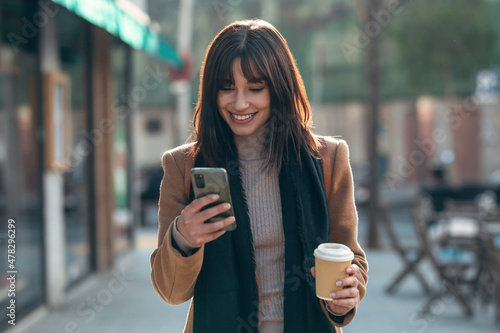 Pretty young woman using her mobile phone while drinking cup of coffee walking the streets of the city Fototapet