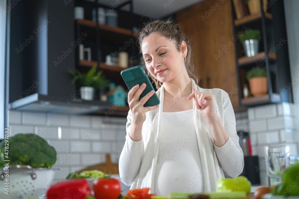 Young pregnant woman preparing healthy food with lots of vegetables at home and use a smartphone to look for some recipes. Healthy diet Young pregnant woman preparing healthy food with lots of vegetables at home and use a smartphone to look for some recipes. Healthy diet