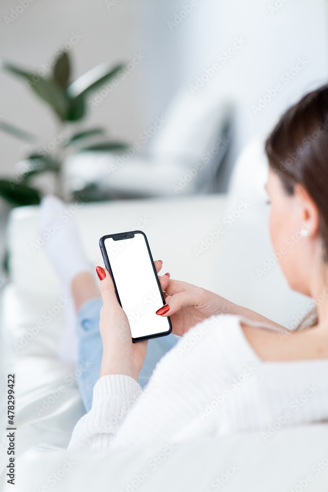 Young woman with modern smartphone lies on couch. Background of light and cozy interior. Phone screen mockup for your advertisement. Young woman with modern smartphone lies on couch. Background of light and cozy interior. Phone screen mockup for your advertisement.