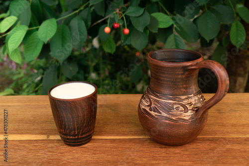 White milk in a clay glass from a jug on a wooden table against the background of green branches of a cherry in the garden