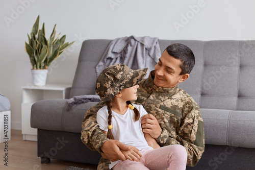 Photos Horizontal shot of satisfied Caucasian soldier man wearing camouflage uniform returning home after army, enjoying time with his little daughter, looking at each other with gentle and love