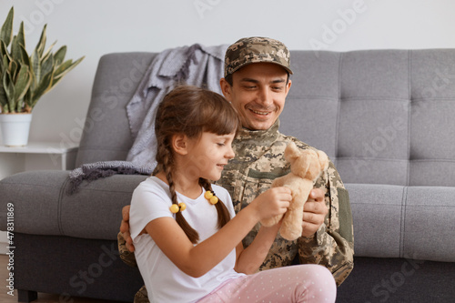 Horizontal shot of delighted father with his kid, Caucasian soldier man wearing camouflage uniform returning home after army, enjoying time with his little daughter, child showing her toy to dad. photo