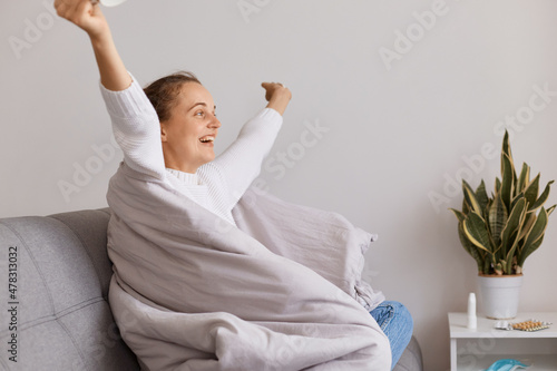Fotografering Indoor shot of happy cute girl sitting on sofa and enjoy about getting better after an illness, recovery after disease, cold or flu, healthcare, satisfied female with raised hands